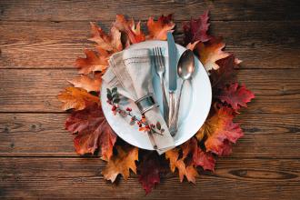 Top view of white plate with napkin and silverware on top of ring of fall leaves on wooden table