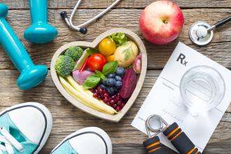 Healthy lifestyle concept with heart shaped bowl with fruits in vegetables surround by shows, weights, glass of water, and prescription pad.