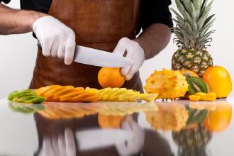 Zoomed in image of person slicing orange with food safe gloves. Sliced limes, oranges, and lemons in front of cutting board.